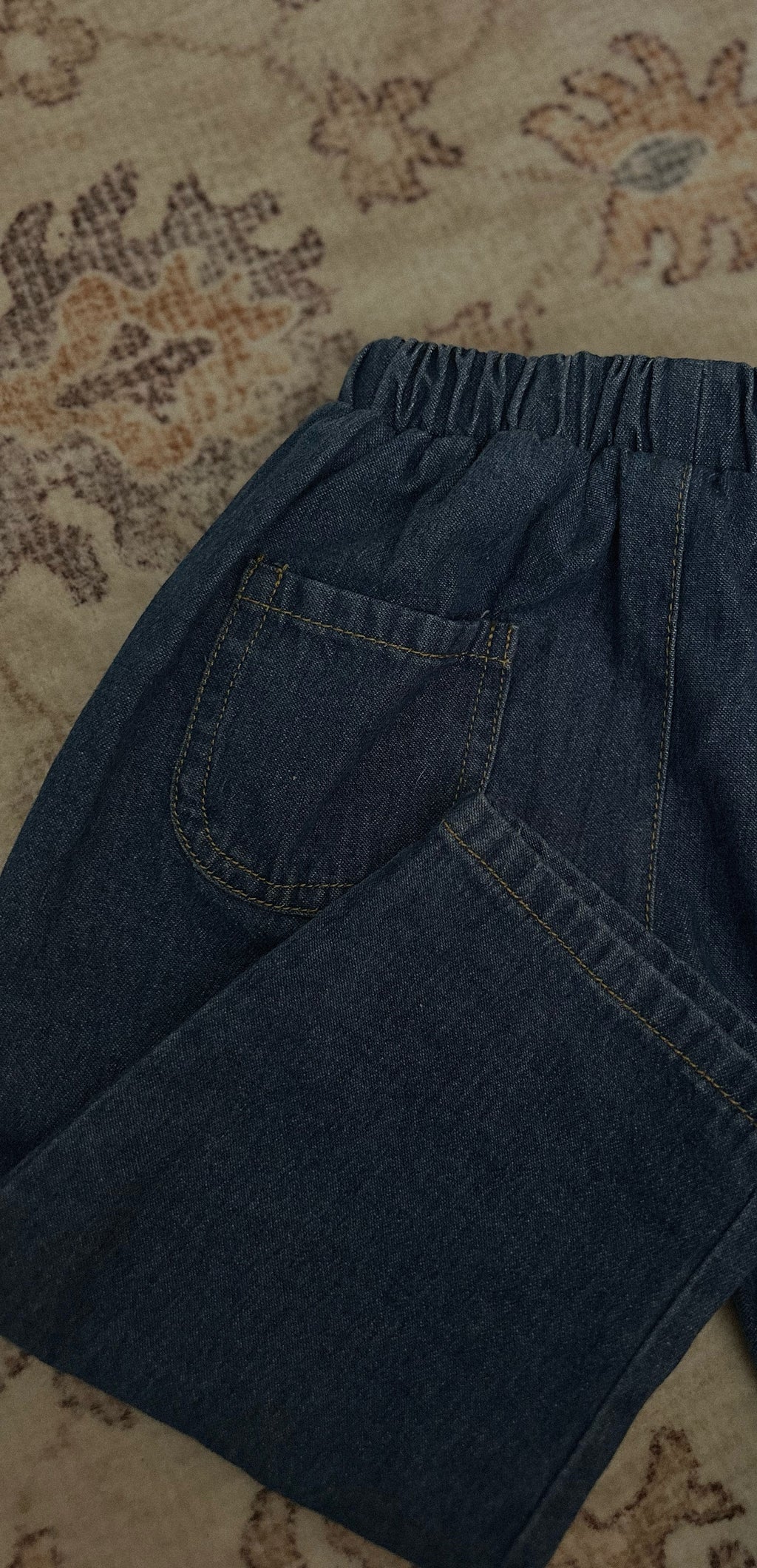 Back of 100% cotton medium to dark wash denim jean laid out on rug. 