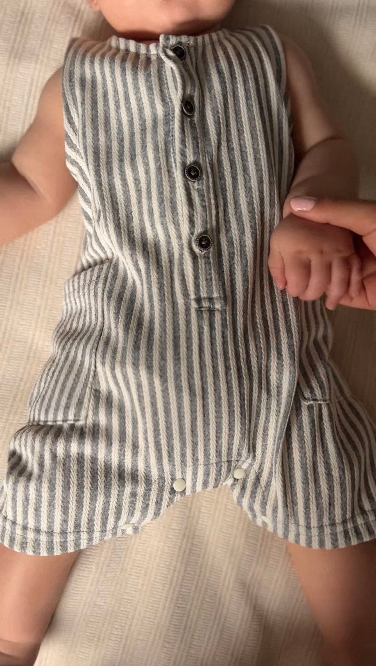 Baby boy wearing a blue and white pinstriped romper jumpsuit with buttons, sitting on a beige surface.